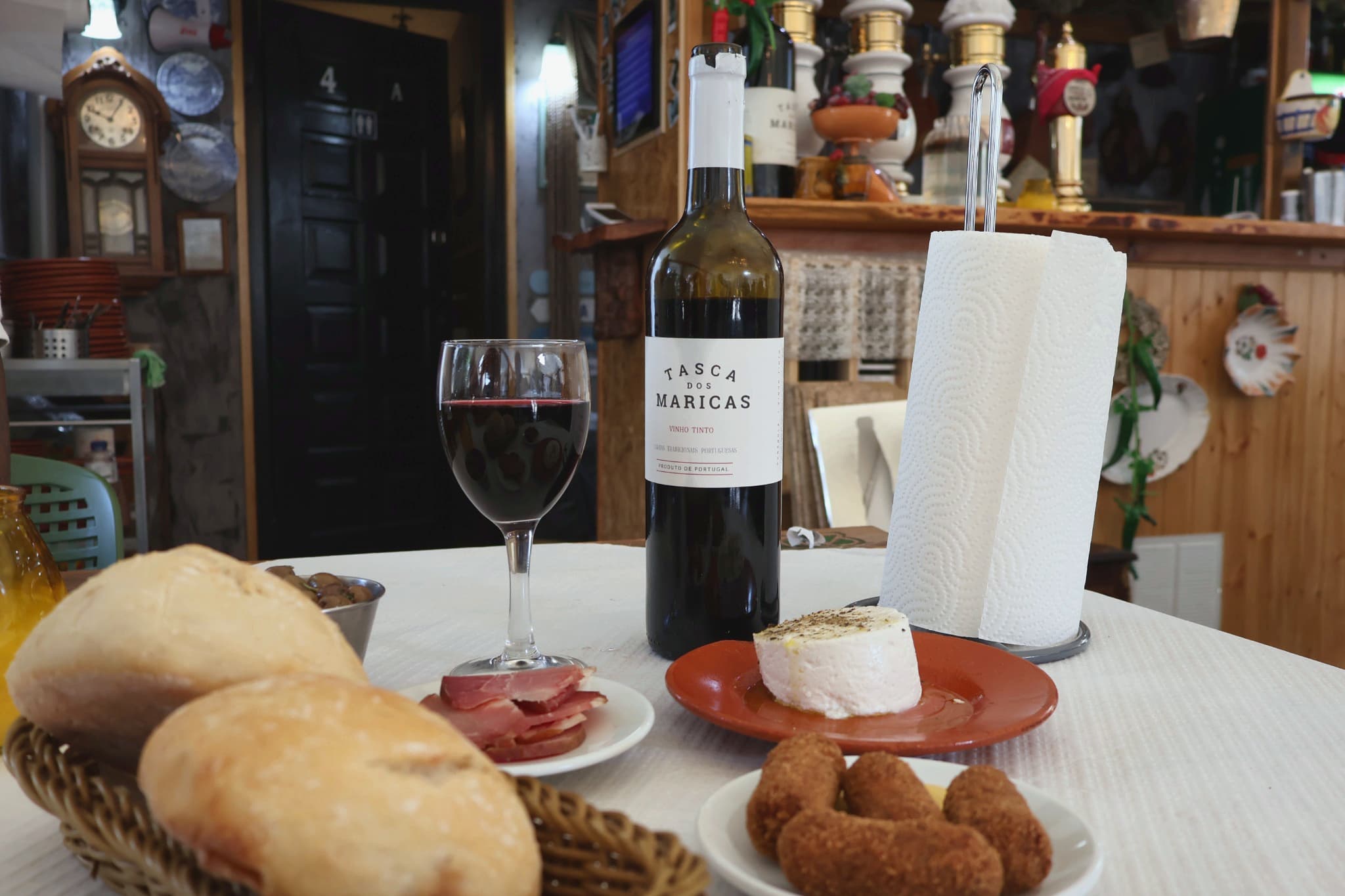 Mesa com pão, presunto, queijo, croquetes e uma garrafa de vinho tinto Tasca dos Maricas, com o balcão ao fundo.